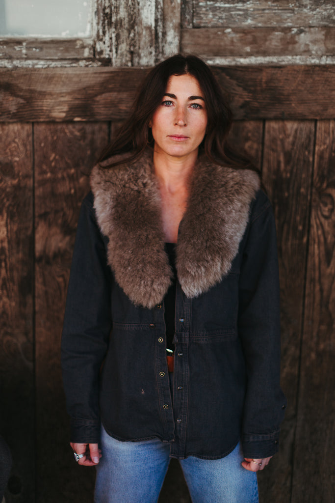 Woman wearing a dark jacket with a fur collar against a wooden wall.