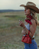 A woman in a cowboy hat and denim jeans stands outdoors in a field, wearing a red bandana, a white tank top, and carrying a small red handbag.