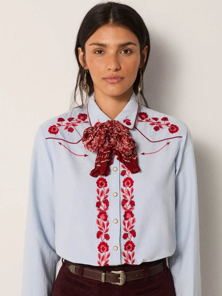 Woman wearing a light blue shirt with red floral embroidery and a matching red scarf against a plain background